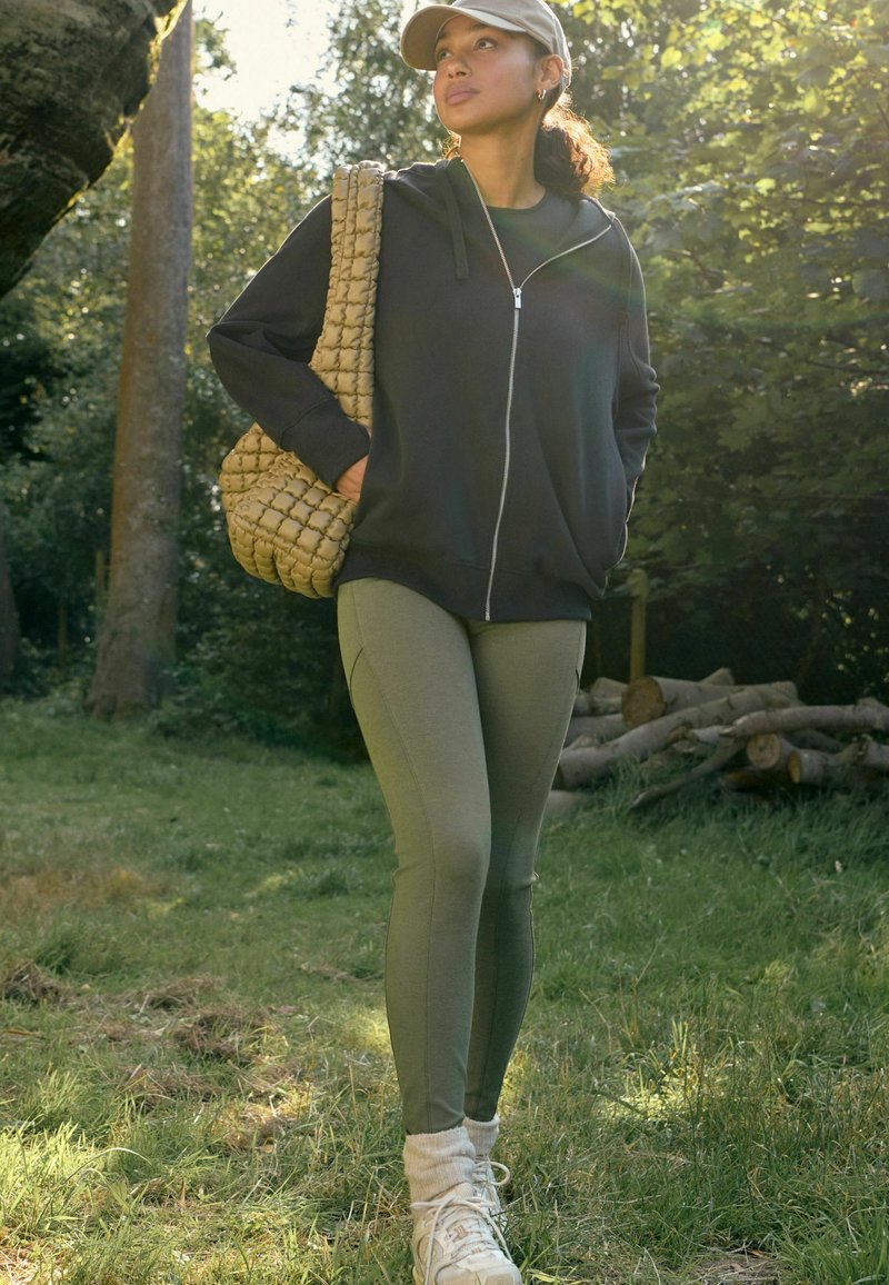 Sudadera negra con cremallera, leggings verdes y zapatillas beigas. La modelo lleva una bolsa de mano de tejido beige y un gorro, de pie en un entorno de césped.