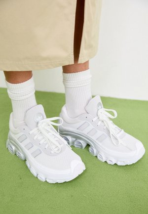 Stopy w białych butach sportowych Adidas z grubymi, profilowanymi podeszwami oraz białych prążkowanych skarpetach, stojące na zielonym dywanie obok beżowej tkaniny.