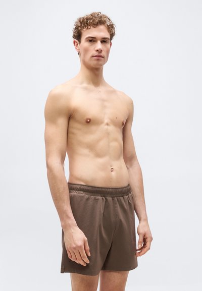 Νέος άντρας με σγουρά μαλλιά που φοράει καφέ σορτς με ελαστική μέση, στέκεται μπροστά σε απλό ανοιχτό γκρι φόντο.