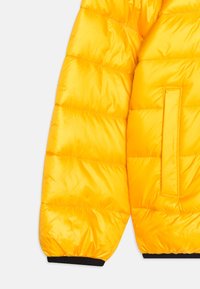 Manga de abrigo acolchado amarillo con una textura suave y brillante. Presenta costuras horizontales y un bolsillo rectangular integrado en el lateral.