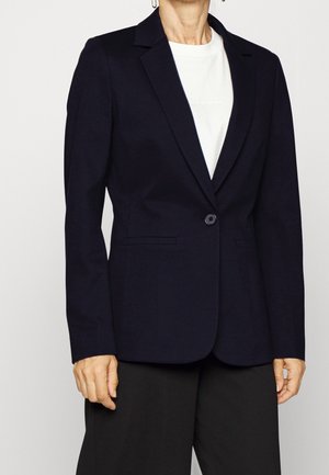 Femme portant un blazer bleu marine sombre ajusté avec un bouton, sur un haut blanc et un pantalon noir, debout devant un fond uni.