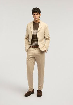 Mand iført beige jakkesæt med brun og beige stribet skjorte, brun bælte og brune loafers, stående mod en ensfarvet baggrund.