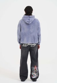 Persona que lleva una sudadera azul desgastada y pantalones vaqueros de pierna ancha negros, con un diseño de script "Ed Hardy" y un colorido tatuaje de calavera en la parte de atrás.