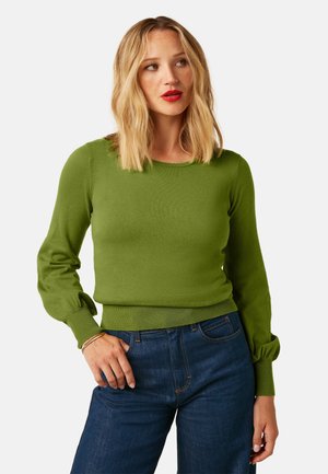Vrouw met schouderlang blond haar, gekleed in een groene trui met lange mouwen en blauwe spijkerbroek, kijkt iets naar de zijkant.