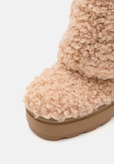 Pehmeä, vaaleanbeige nilkkuri, jossa on kihara pinta ja paksu ruskea pohja, pyöreä kärki ja pehmeä, pörröinen materiaali.