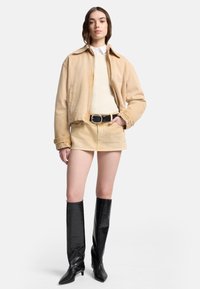 Beżowa jeansowa kurtka na kremowym swetrze, zestawiona z beżową jeansową mini spódnicą i błyszczącymi czarnymi, sięgającymi do kolan, spiczastymi kozakami. Gładka faktura.