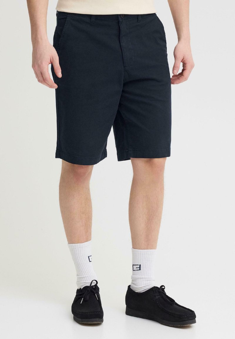 Homme portant un short noir mi-cuisses, des chaussettes blanches montantes avec un logo, et des chaussures noires à lacets, debout devant un fond uni.
