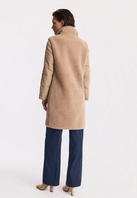 Beżowy płaszcz z owczej wełny z wysokim kołnierzem, beżowe podszyte rękawy, w połączeniu z ciemnoniebieskimi spodniami z szerokimi nogawkami i jasnobeżowymi obcasami.