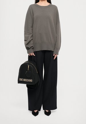 Personne portant un pull gris ample et un pantalon large noir tenant un sac à dos noir Love Moschino avec des fermetures éclair dorées.