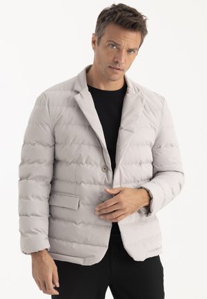 Homme portant un blazer matelassé gris clair sur une chemise noire, ajustant le bouton devant, debout devant un fond blanc uni.