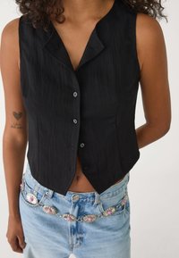 Czarny bezrękawnik z guzikami i dekoltem w kształcie litery V, uszyty z teksturowanej tkaniny, w połączeniu z jasnoniebieskimi spodniami typu high-waisted i srebrnym paskiem w kwiaty.