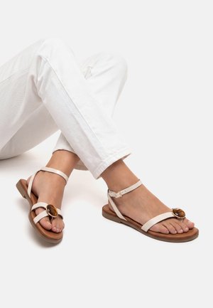 Piedi che indossano sandali piatti bianchi con cinturini alla caviglia e grandi decorazioni circolari marroni, abbinati a pantaloni bianchi.