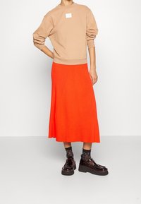 Sudadera color beige con cuello redondo y pequeño parche con logo, combinada con falda midi naranja acanalada y zapatos negros de plataforma con acabado brillante.