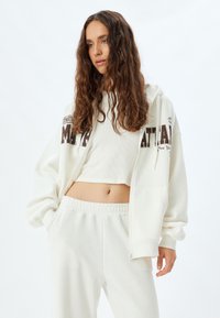 Weiße Zip-Hoodie mit braunem "New York"-Schriftzug und Logodetails, kombiniert mit einem weißen gerippten Crop-Top und passenden Jogginghosen. Weicher Stoff.