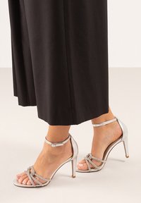 Sandalias de tacón alto plateadas con tiras finas, punta abierta y hebilla en el tobillo. Combinadas con pantalones anchos negros y fluidos. Textura suave.