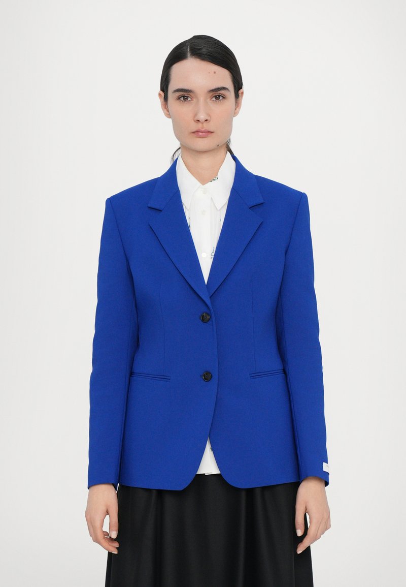 Bright blue tailored blazer with structured shoulders, two black buttons, and side pockets, layered over a white collared shirt.
