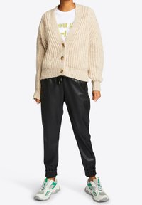 Beżowy sweter z dzianiny z dekoltem w serek, z przodem na guziki i prążkowaną teksturą, w połączeniu z czarnymi joggerami z sztucznej skóry i szaro-zielonymi trampkami.