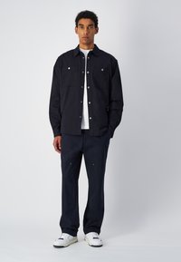 Czarna koszula zapinana na guziki z dwiema kieszeniami na piersi, zestawiona z ciemnymi spodniami z zamkami błyskawicznymi oraz białymi sneakersami. Czysty, minimalistyczny design.