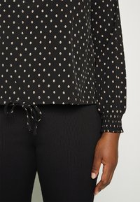 Blouse noire à manches longues avec un petit motif floral blanc et un ourlet à cordon, associée à un pantalon noir côtelé, main reposant sur le côté.