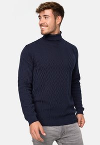 Mørkeblå strikket polo genser med zigzag-mønster, ribbede mansjetter og kant. Materialet ser mykt ut med en teksturert finish.