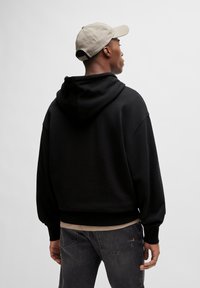 Zwarte hoodie met een relaxte pasvorm, geribbelde manchetten en zoom, gecombineerd met een lichtbeige shirt en donkere jeans. Vergezeld van een beige pet.