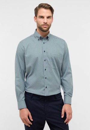 Geruit groen en marineblauw button-up shirt met een kraag, lange mouwen en marineblauwe knopen. Gedragen met donkere broeken, die een aansluitend ontwerp tonen.