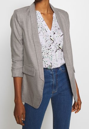 Personne portant un blazer gris sur un chemisier fleuri avec des imprimés d'oiseaux, associé à un jean taille haute bleu, sur un fond uni.