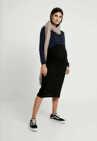 Conjunto de maternidade com uma blusa de manga longa azul-marinho, uma saia preta canelada até os joelhos e ténis pretos. Um cachecol bege envolve o pescoço.