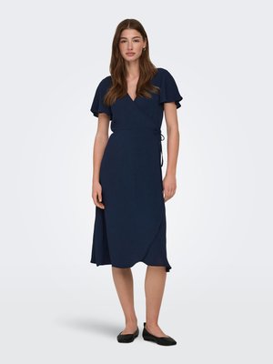 Jeune femme debout, portant une robe portefeuille bleu marine longueur genou avec des manches volantées et des chaussures plates noires, sur un fond clair uni.