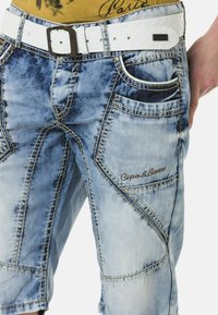 Blaue Jeanshose mit einem verblassten Look, kontrastierenden Nähten, mehreren Taschen und einem weißen Gürtel mit einer silbernen Schnalle. Logo sichtbar.