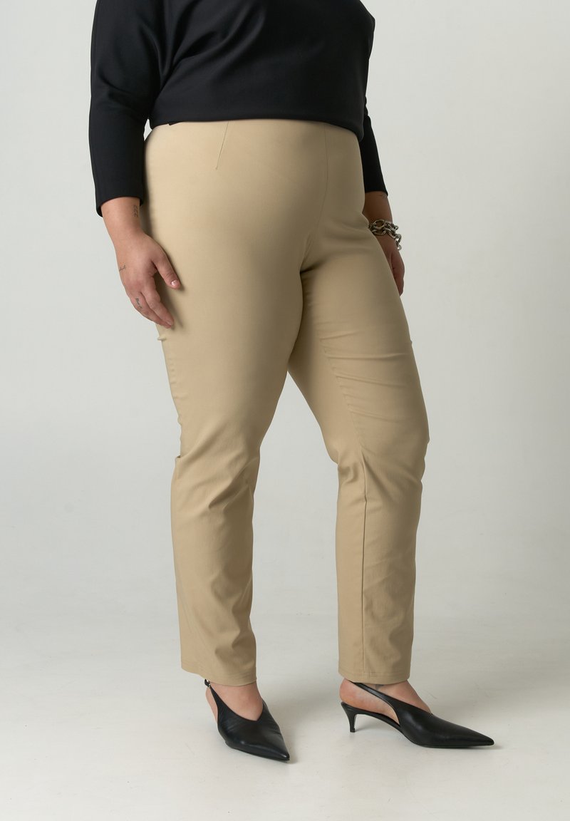 Beige nauwsluitende broek met een gladde textuur, gecombineerd met zwarte schoenen met puntige neus. Zwart topje met driekwart mouwen maakt de outfit compleet.