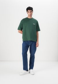 Tricou din bumbac verde, cu mâneci scurte și guler rotund, asortat cu blugi din denim albastru. Modelul poartă adidași albi, stând în fața unui fundal alb.