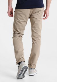 Pantalons en coton beige avec une coupe slim, présentant une texture lisse et un design classique à cinq poches, associés à des chaussures de sport gris clair.