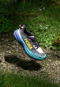 HOKA running shoe with a beige and black upper, colorful accents in blue and purple, featuring a textured sole and distinctive branding.
