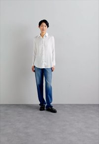 Camicia bianca con bottoni e maniche lunghe, abbinata a jeans in denim blu chiaro. La persona indossa scarpe nere e si trova di fronte a uno sfondo grigio.