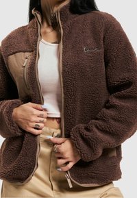 Giacca in pile marrone con zip davanti, caratterizzata da un pannello beige, orlo a costine e logo ricamato. Texture morbida, maniche lunghe e vestibilità rilassata.