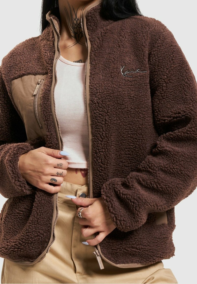 Giacca in pile marrone con zip davanti, caratterizzata da un pannello beige, orlo a costine e logo ricamato. Texture morbida, maniche lunghe e vestibilità rilassata.