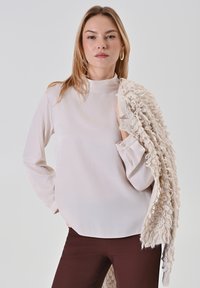 Helles Beige, langärmliges Oberteil mit hohem Kragen, kombiniert mit einem strukturierten, cremefarbenen Schal im Boucle-Design. Braune Hose.
