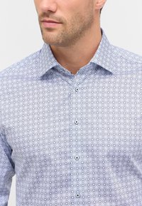Camicia a maniche lunghe con fantasia che presenta una base azzurra chiara con disegni circolari in blu scuro, e un colletto classico con dettagli di bottoni.