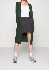 Grön lång kofta med öppen framsida och sidofickor, lager på lager över en vit tanktop och svart randig kjol. Silverfärgade sportskor med vita strumpor.