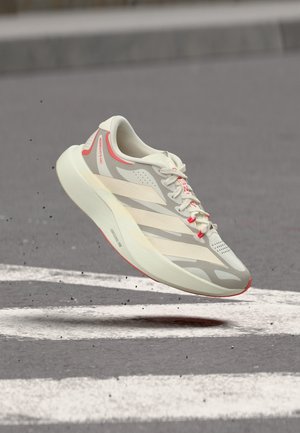 Világosszürke és fehér futócipő piros díszítésekkel, fehér vonallal jelölt járda felett lebegve, mutatva a talp és az oldal részleteit.