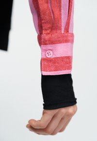 Χέρι που φοράει μανσέτα πουκάμισου με μακριά μανίκια, κόκκινη με ροζ ρίγα, με ροζ κουμπί πάνω σε μαύρο ριμπ μανίκι, σε λευκό φόντο.