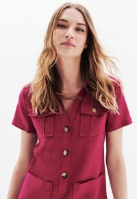 Abito fucsia con bottoni, maniche corte, tasche sul petto e due tasche anteriori. Realizzato in tessuto texturizzato, con bottoni rotondi.