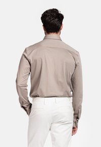 Beige blouse met lange mouwen en een klassieke kraag, met een gladde textuur en knopenmanchetten, gecombineerd met witte broek, gezien vanuit de achterkant.