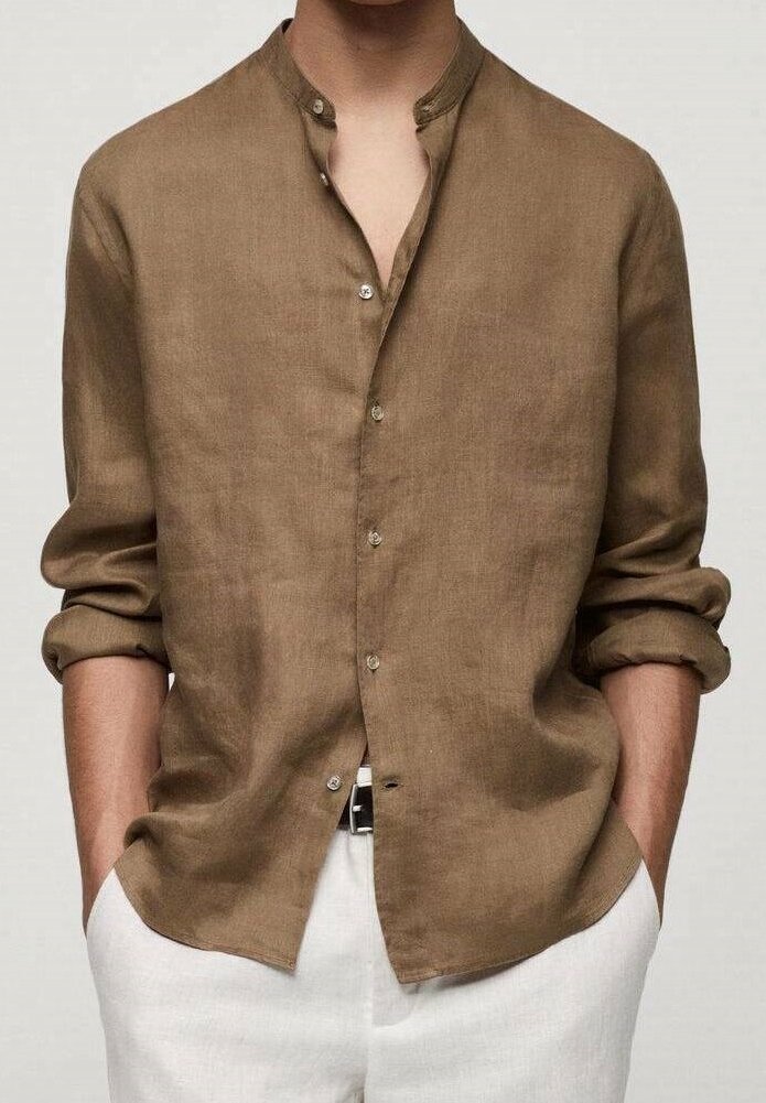 Chemise en lin marron à manches longues avec col mao et manches retroussées, portée non rentrée sur un pantalon blanc, les mains dans les poches.