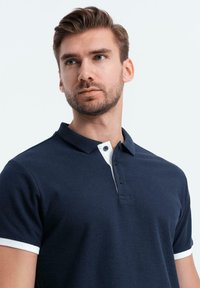 Námořnická modrá polo košile s bílým límečkem a manžetami, vyrobená z texturované bavlny. Má tříknoflíkovou légou a krátké rukávy.