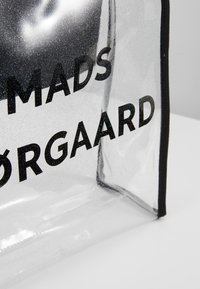 Sac clair et transparent avec bordure noire et texte noir indiquant "MADS ØRGAARD" sur une surface blanche.