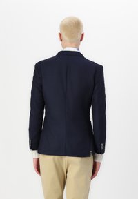 Blazer blu navy con taglio strutturato, spacco singolo e polsini abbottonati. Indossato sopra una camicia a motivi chiari e abbinato a pantaloni beige.