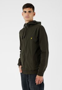 Giacca verde oliva con zip e cappuccio, dotata di un piccolo logo giallo sul petto, realizzata in tessuto liscio e progettata con tasche laterali.