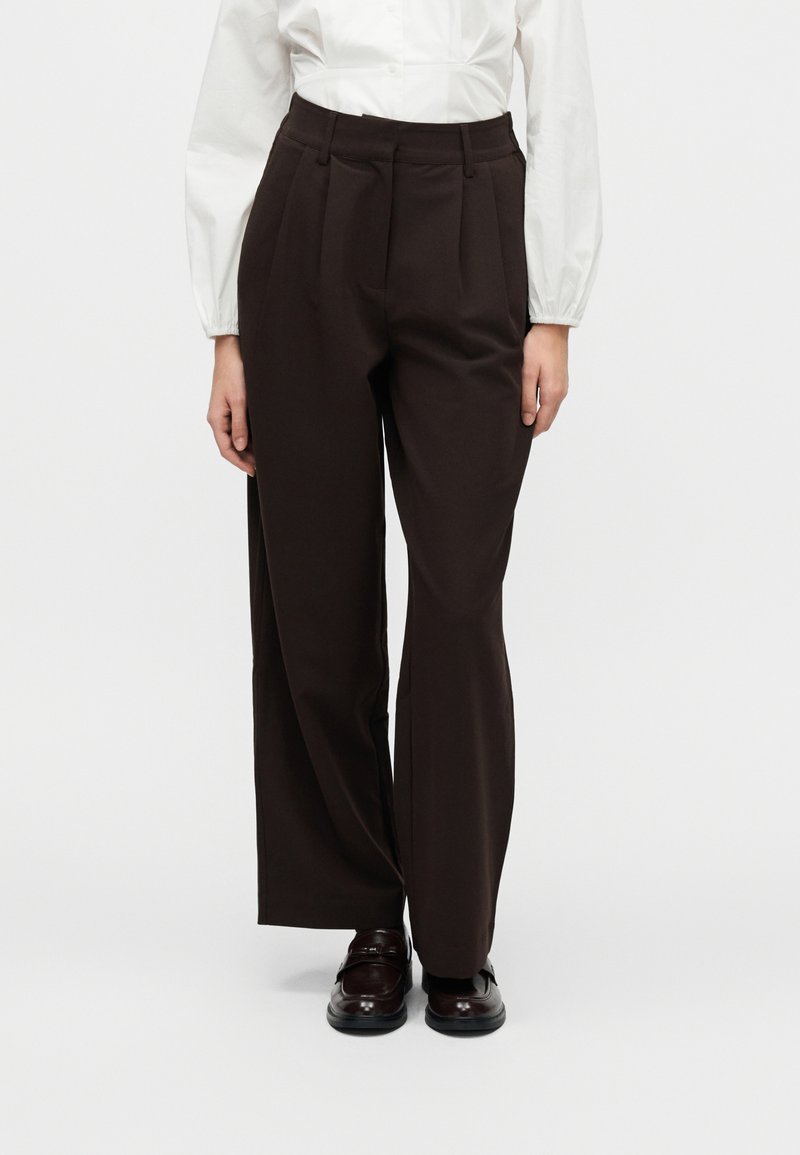 Καφέ παντελόνι με ψηλή μέση από λείο ύφασμα, με πιέτες, ίσια μπατζάκια και πλαϊνές τσέπες. Συνδυασμένο με σκούρα μοκασίνια.